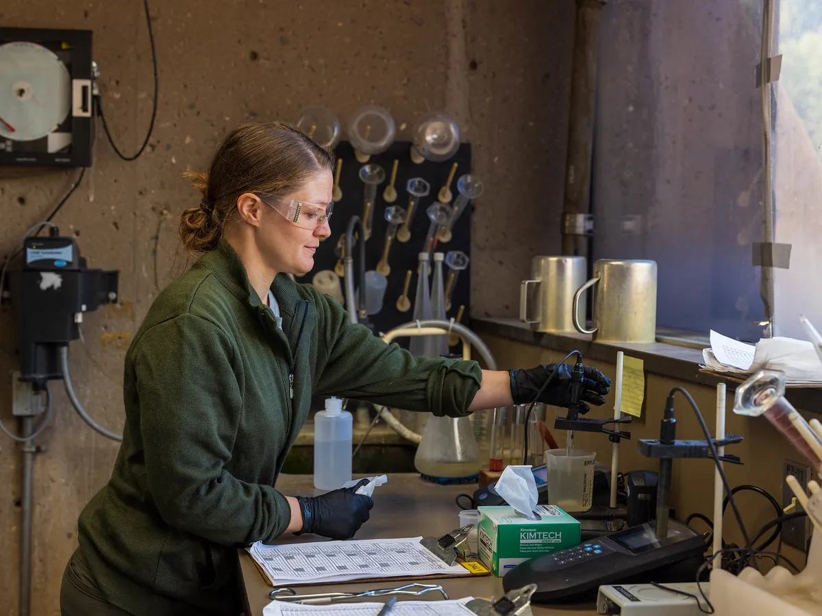 A technician in safety glasses checks a water sample at a test bench lined with sample bottles